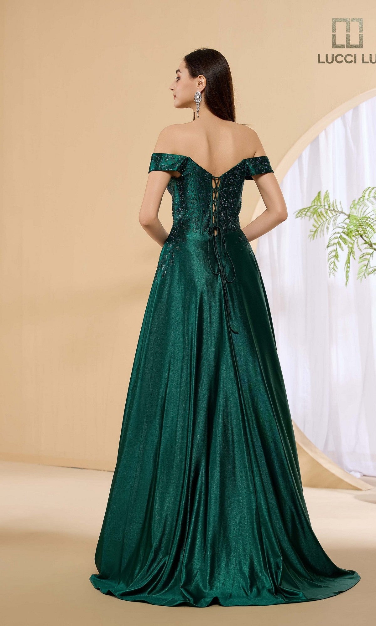 A woman stands facing away in a minimal indoor setting, wearing the Lucci Lu 1483 long prom dress—an emerald green, shiny off-the-shoulder A-line gown with a lace-up corset bodice.